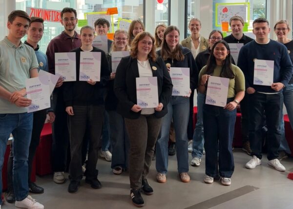 Gruppenfoto mit rund 20 Menschen, die alle je ein Zertifikat in den Händen halten und für ein Gruppenfoto zusammenstehen und in die Kamera gucken.